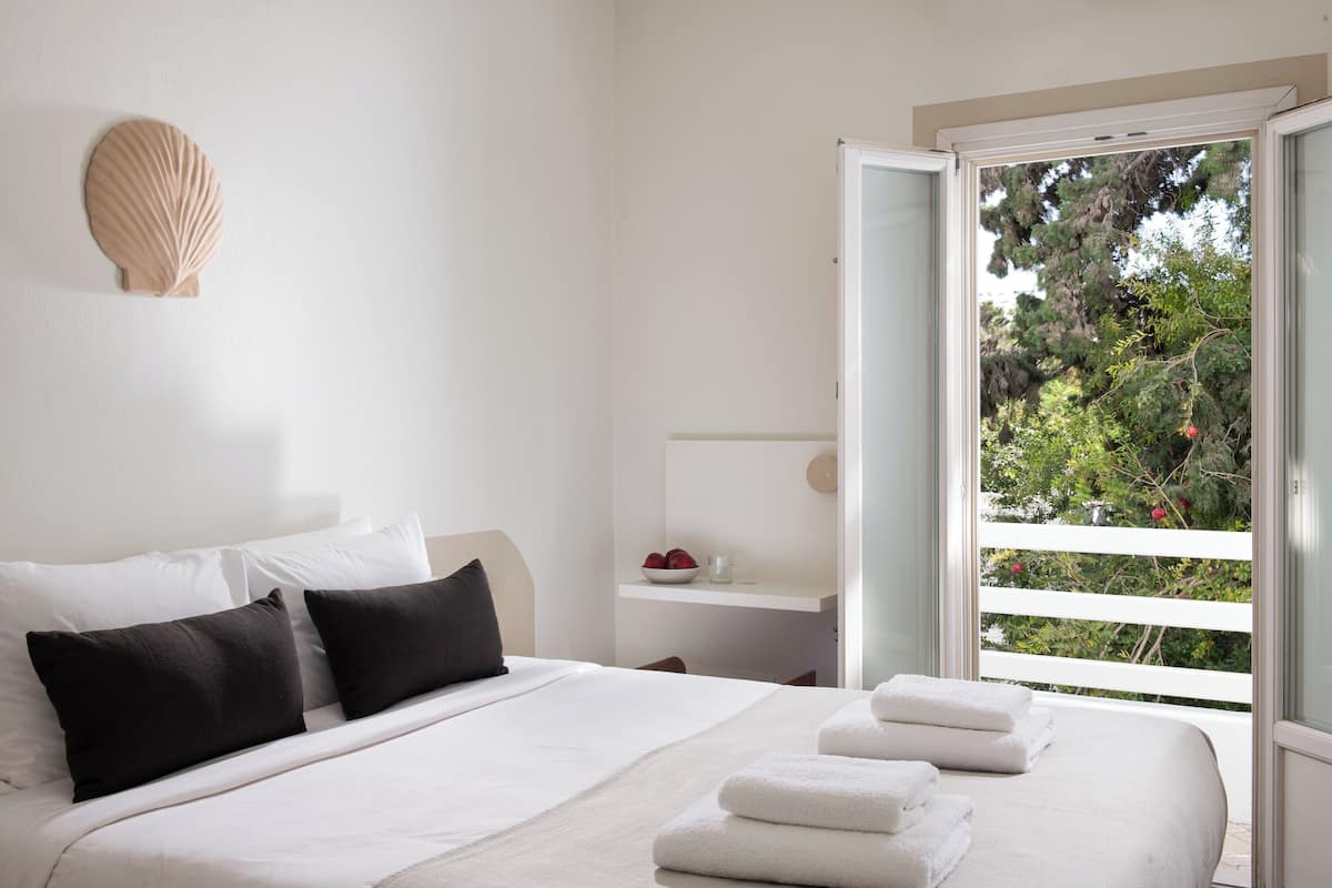 Un dormitorio con cama, almohadas, una mesita de noche, un cuenco, una taza y vistas a árboles a través de una ventana.