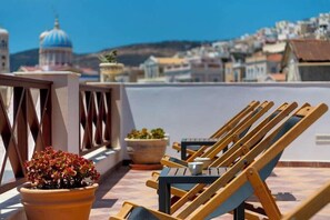 A terrace with wooden lounge chairs, potted plants, and a view of a coastal town with buildings and a blue-domed structure. - Electra (Syros)