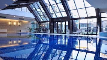 Una piscina interior con techos de vidrio transparente, sillas de descanso y vistas al exterior.