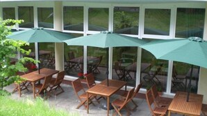 Un patio al aire libre con mesas y sillas de madera, cubierto por sombrillas verdes, y grandes ventanales al fondo.