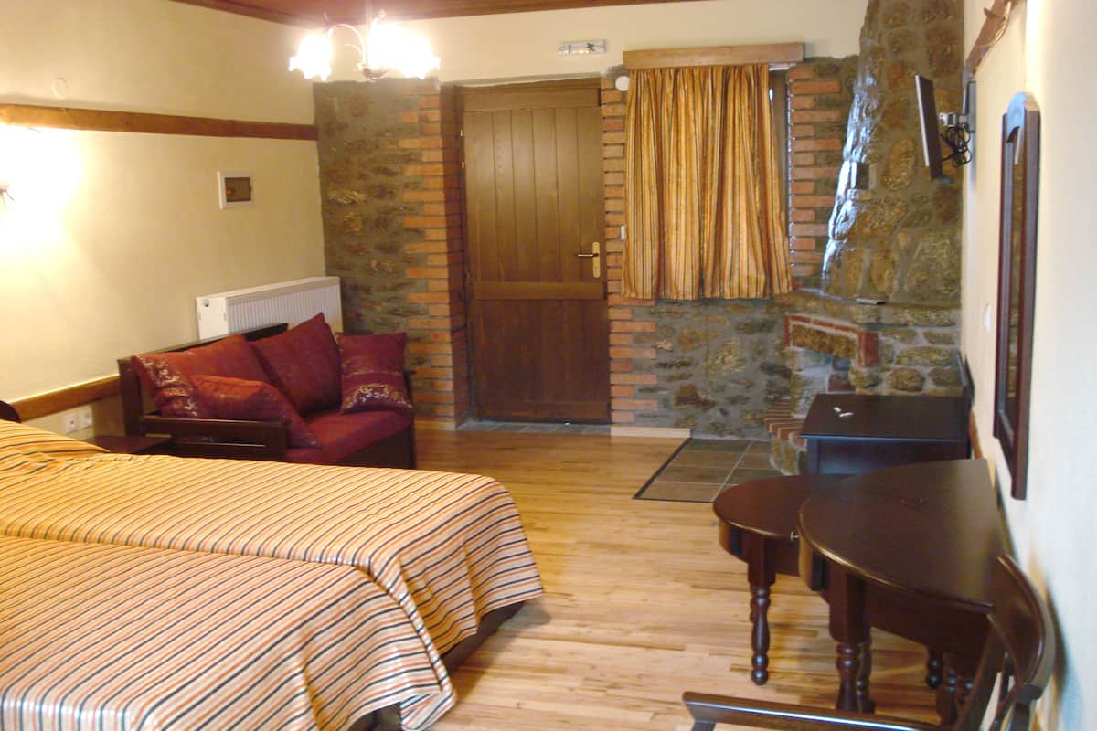 A room with a wooden floor, a stone wall, a wooden door, a red sofa, a wooden table, and a bed with striped bedding.