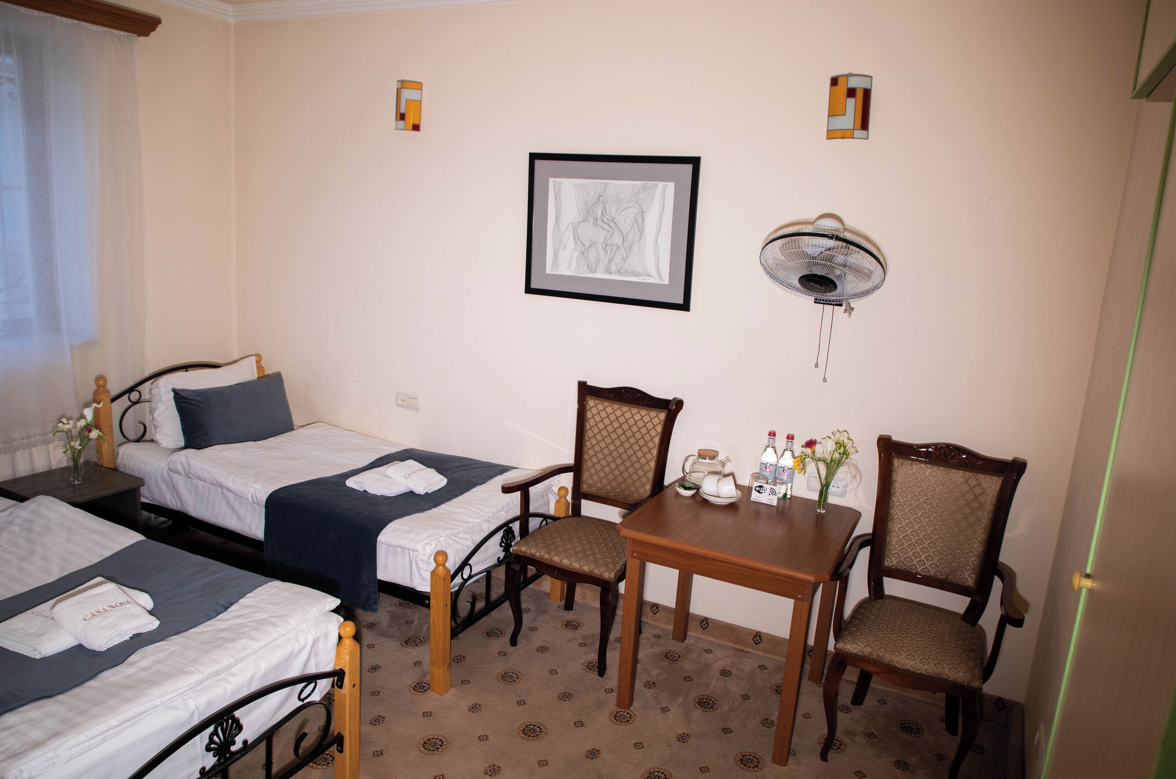A hotel room with two single beds, a wooden table with chairs, a wall-mounted fan, and a framed picture.
