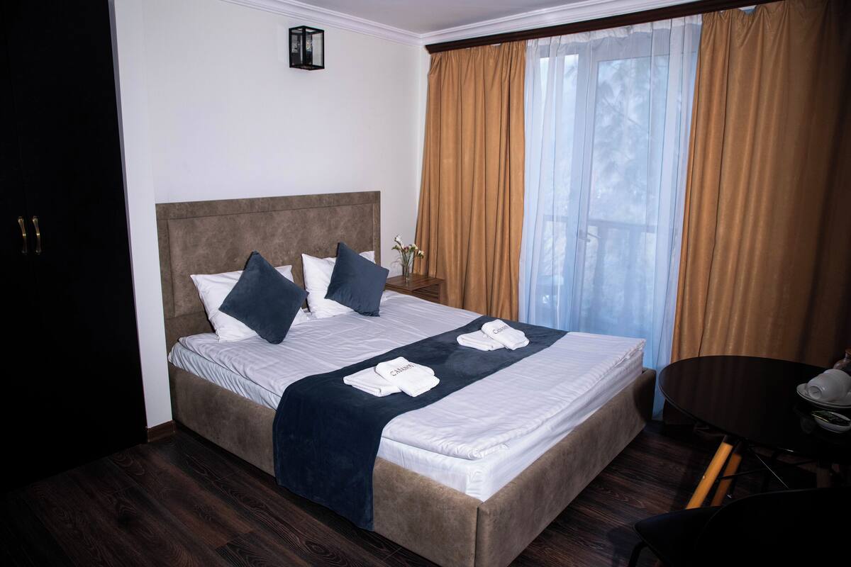 A hotel room with a large bed, a small table, and a view of the outdoors.