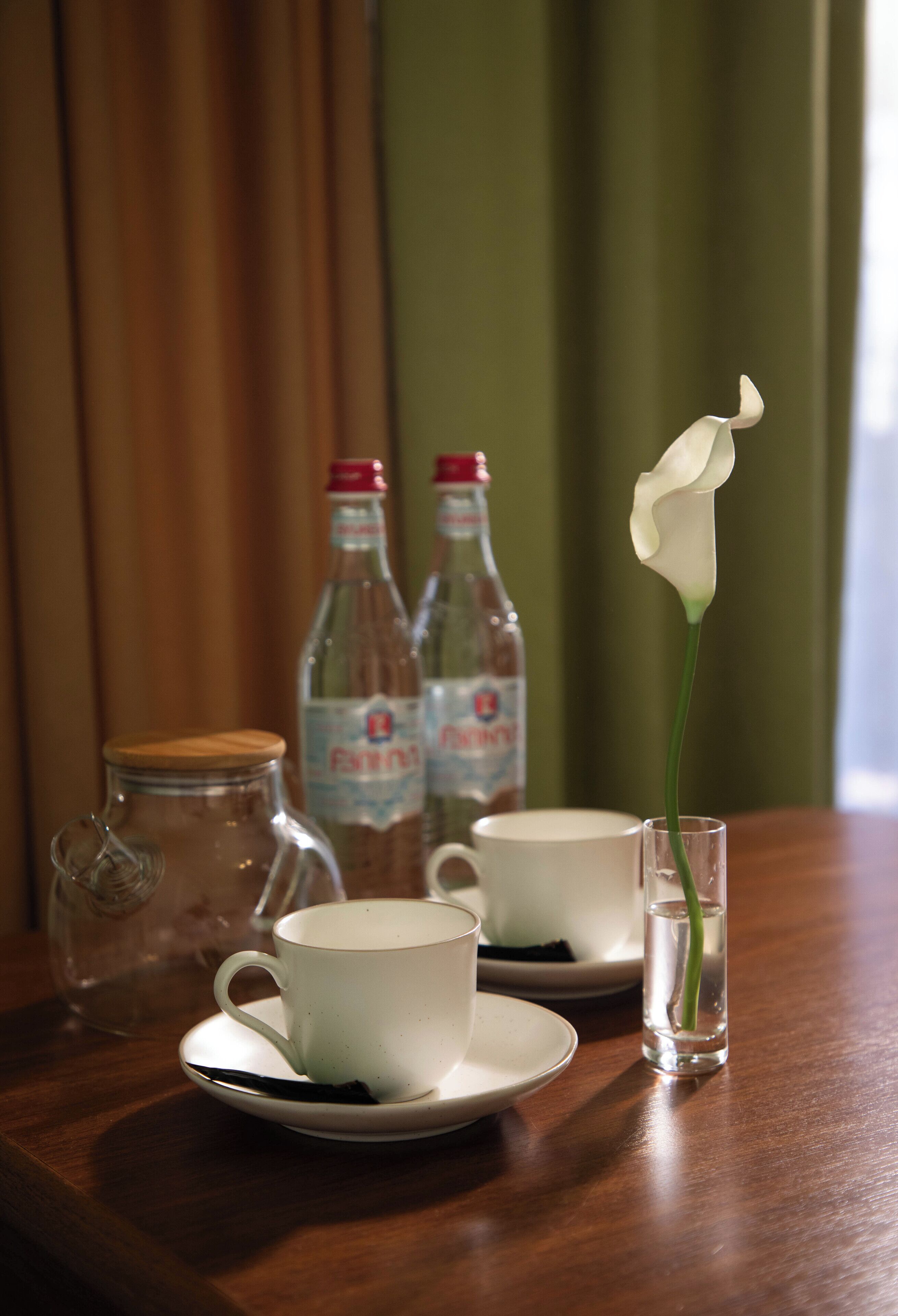 Una mesa con dos tazas blancas, un tarro de vidrio y un florero con una sola flor blanca.