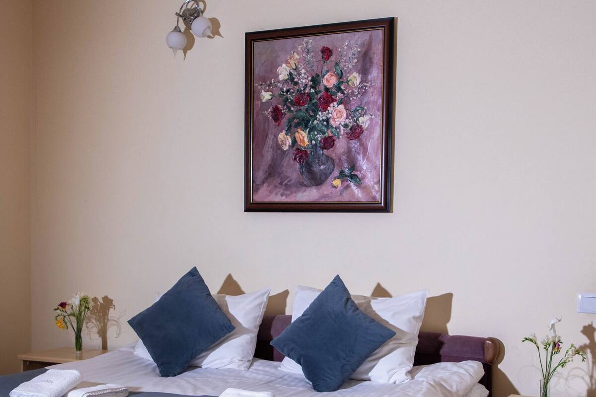 A neatly made bed with a purple headboard, a framed floral painting above it, and a bedside table with a vase of flowers.