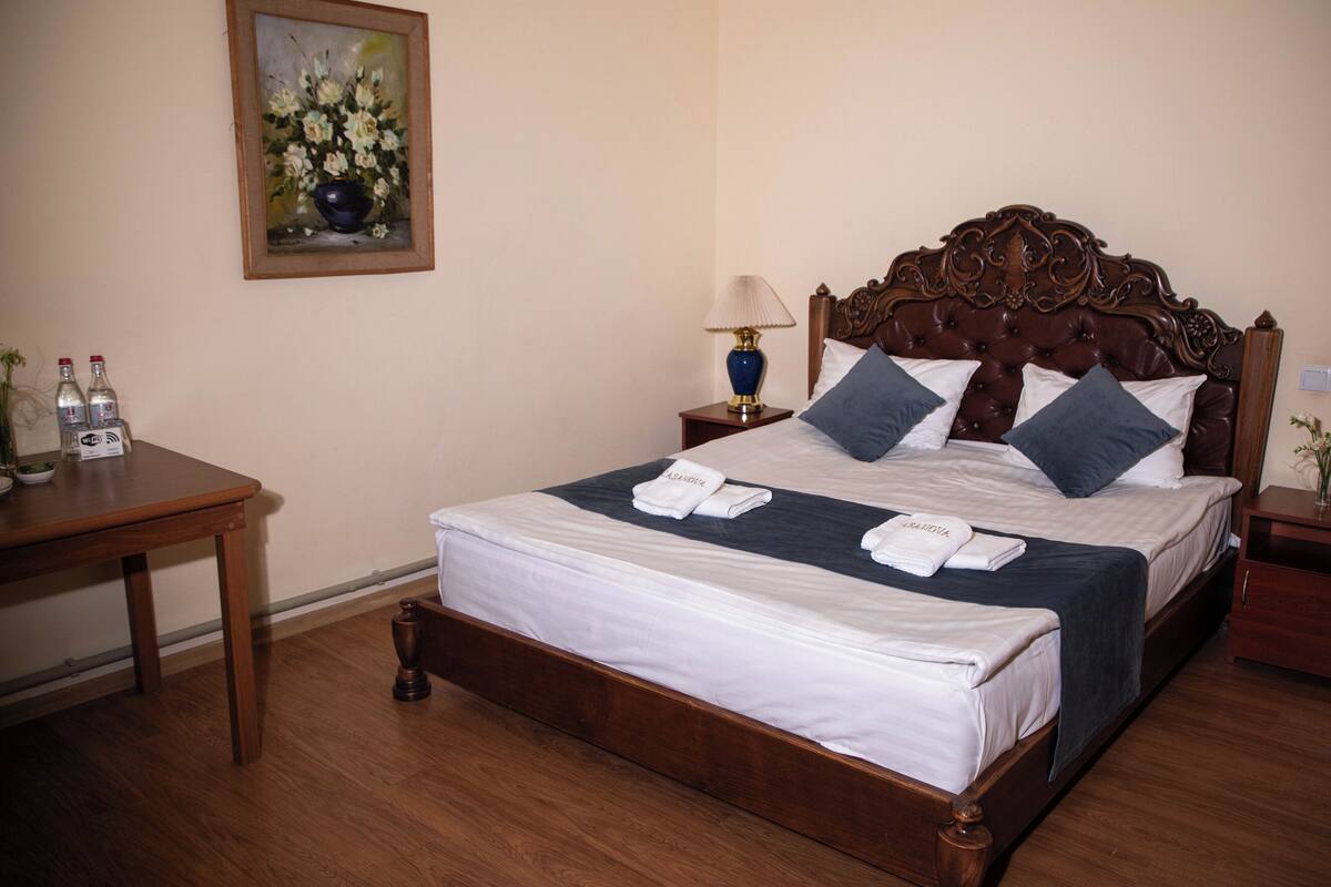 A neatly made bed with white linens and two pillows, a wooden nightstand with a lamp, and a painting on the wall.