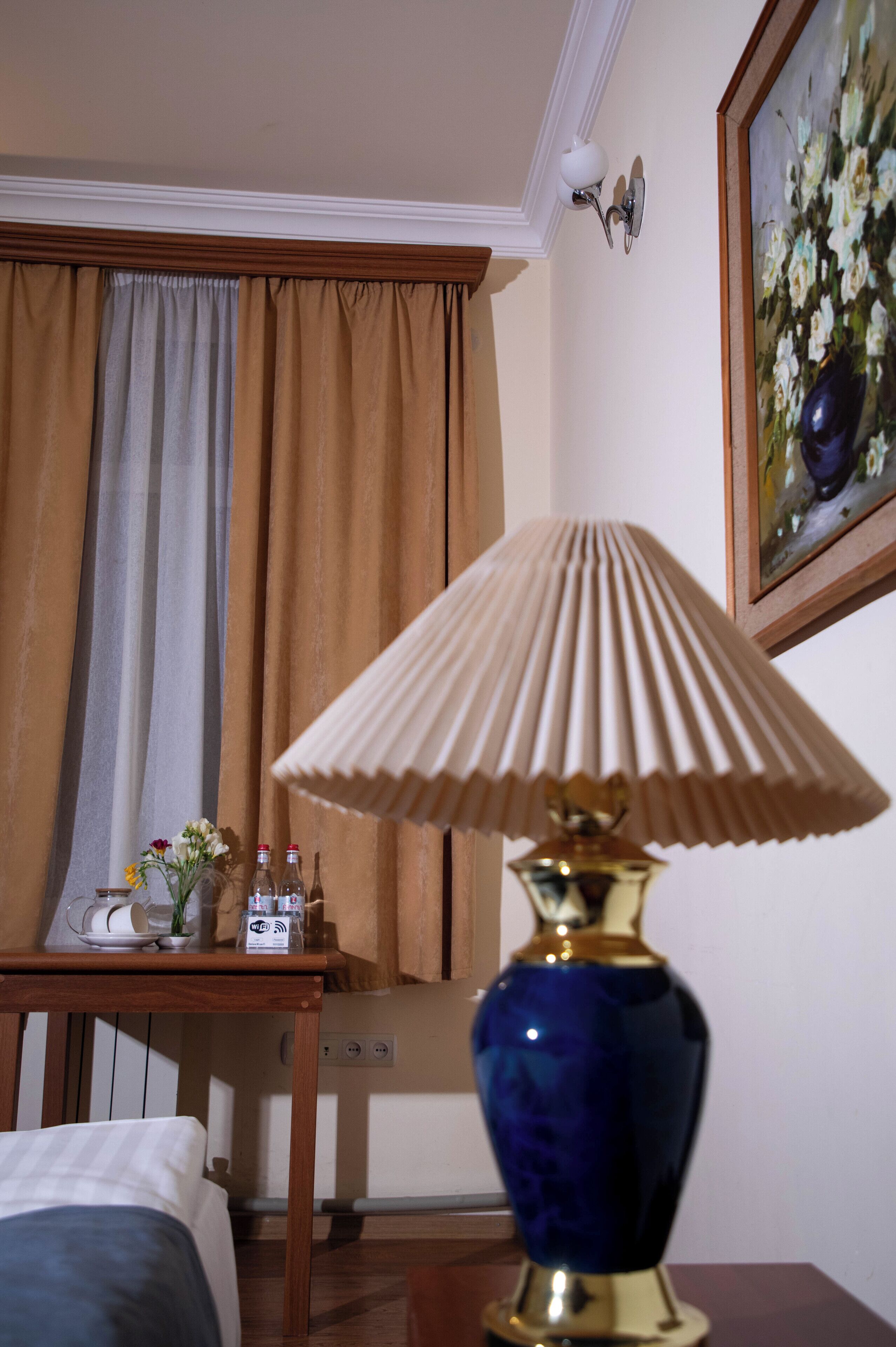 Een hotelkamer met een bed, een tafel met lamp, een schilderij aan de muur en een raam met gordijnen.