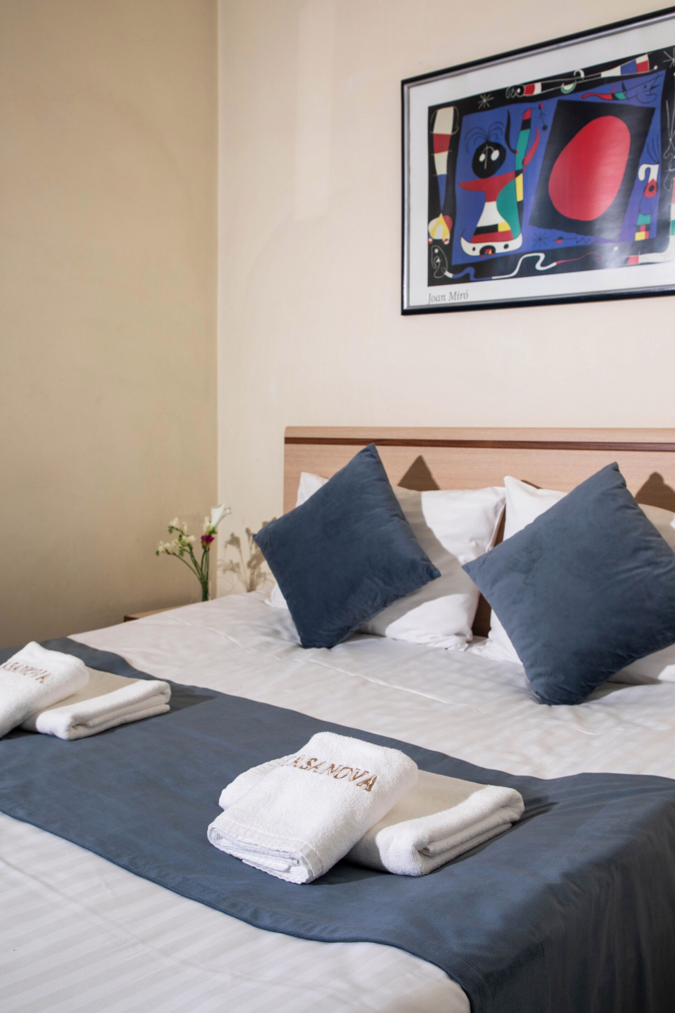 Una cama bien hecha con sábanas blancas, almohadas azules y una colcha azul. Encima de la cama, en la pared, hay un cuadro abstracto enmarcado.