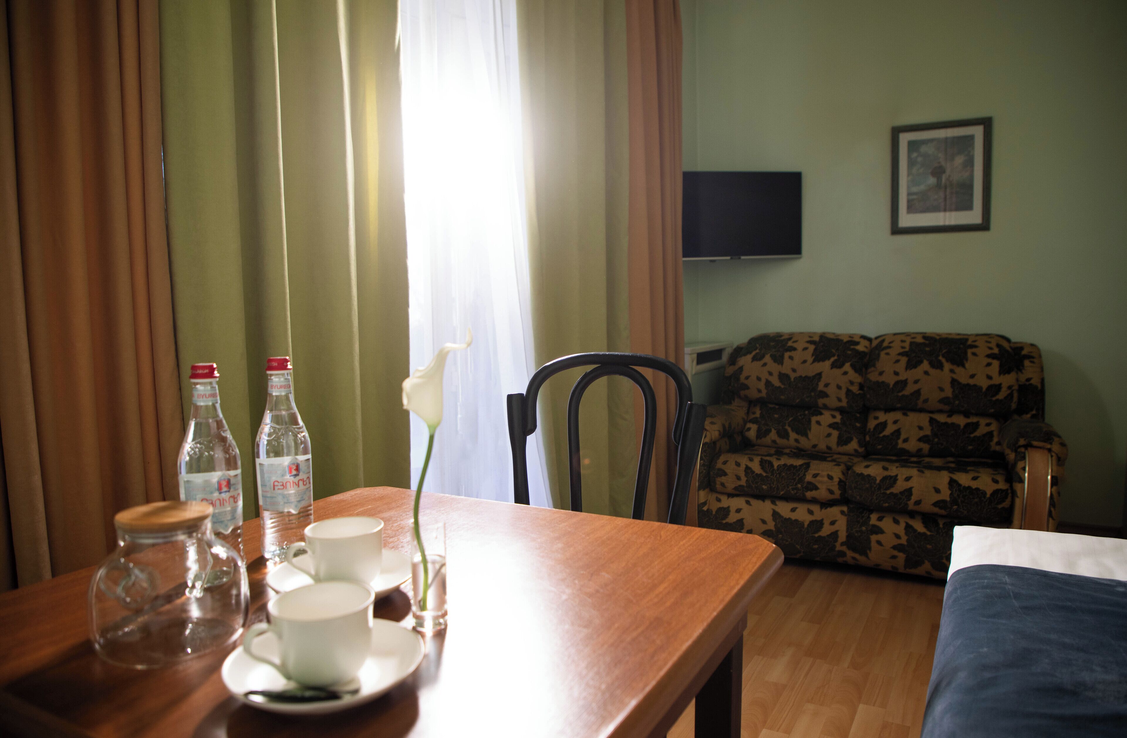 A dining table with a vase of flowers, two bottles of water, and a white rose. A television is mounted on the wall, and a patterned sofa is visible in the background.
