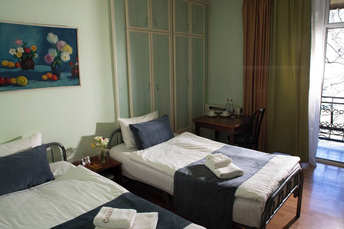 A hotel room with two beds, a painting on the wall, a window with curtains, and a small table with a bottle.