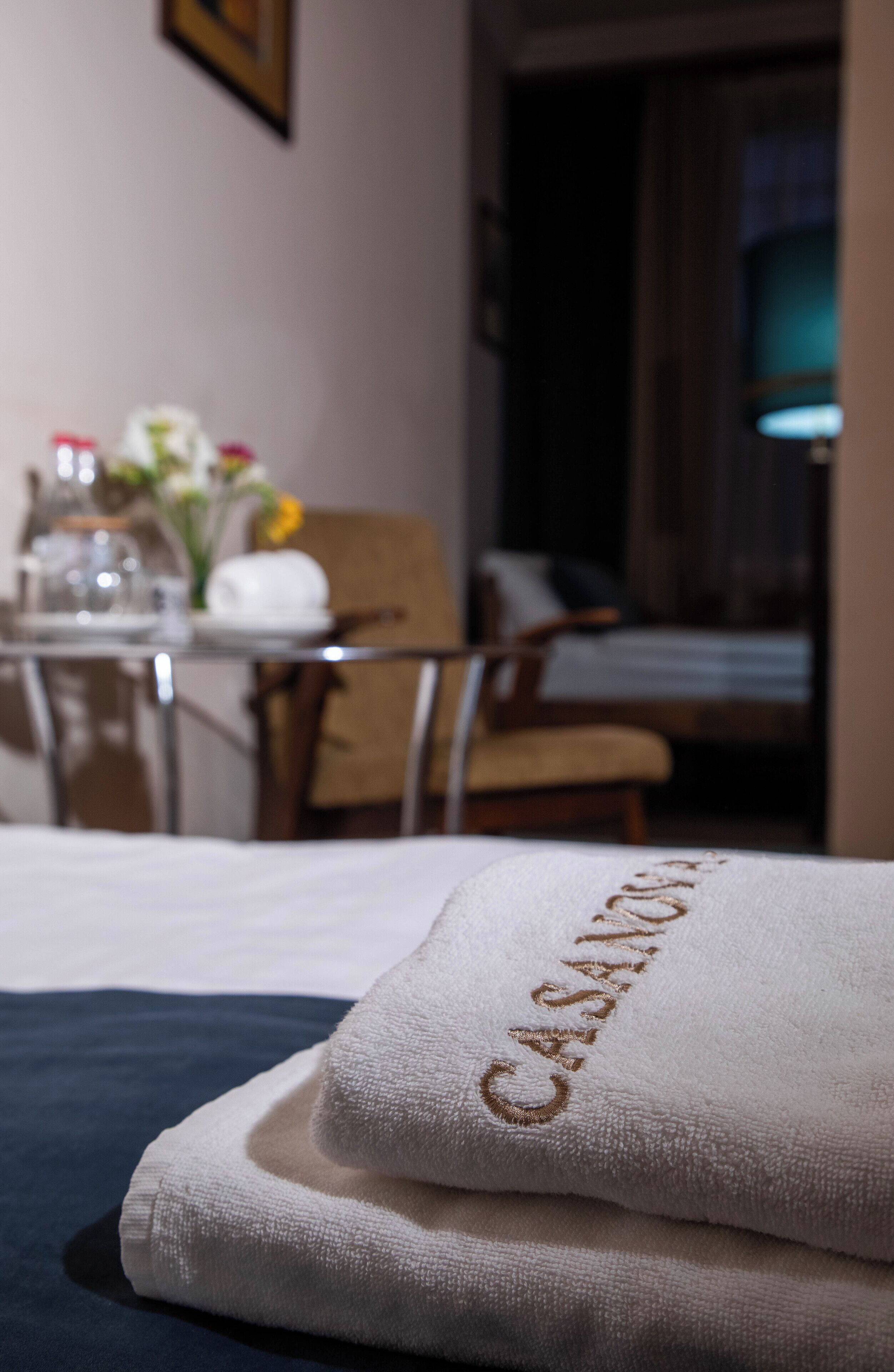Een netjes opgemaakt bed met een witte handdoek met geborduurde tekst, een houten stoel, een kleine tafel met een bloemenvaas, en een kamer met gedempt licht.