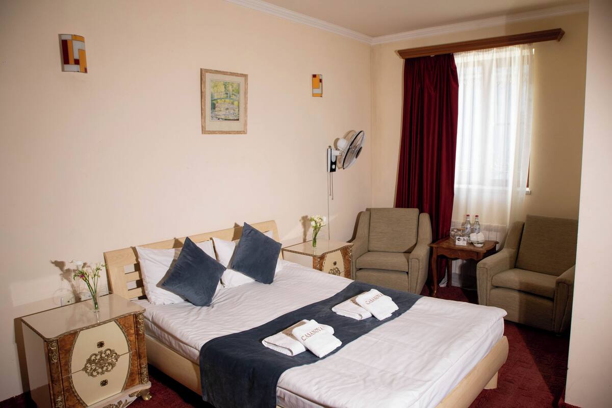 A hotel room with a bed, two armchairs, a small table with a lamp, a bedside table with a vase of flowers, and a window with curtains.