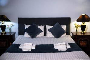Una cama bien tendida con un cabecero oscuro, almohadas blancas y oscuras, y dos toallas con el bordado 'CASANOVA'.