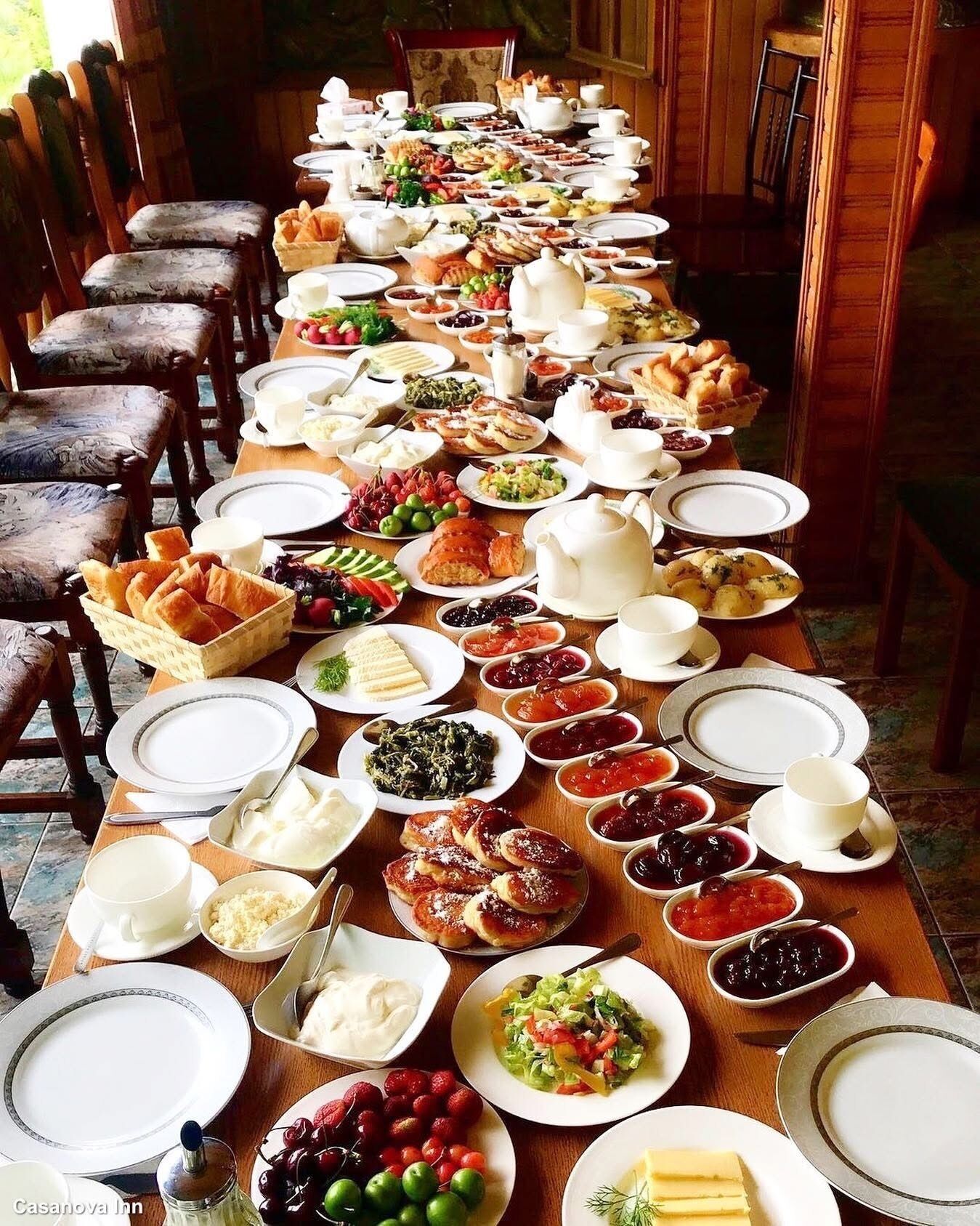 Une longue table à manger, dressée avec divers plats, notamment des fruits, des pâtisseries et des condiments, dans une pièce dont le mobilier et les chaises sont en bois.