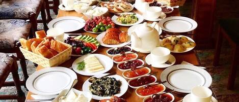 Une longue table à manger, dressée avec divers plats, notamment des fruits, des pâtisseries et des condiments, dans une pièce dont le mobilier et les chaises sont en bois.