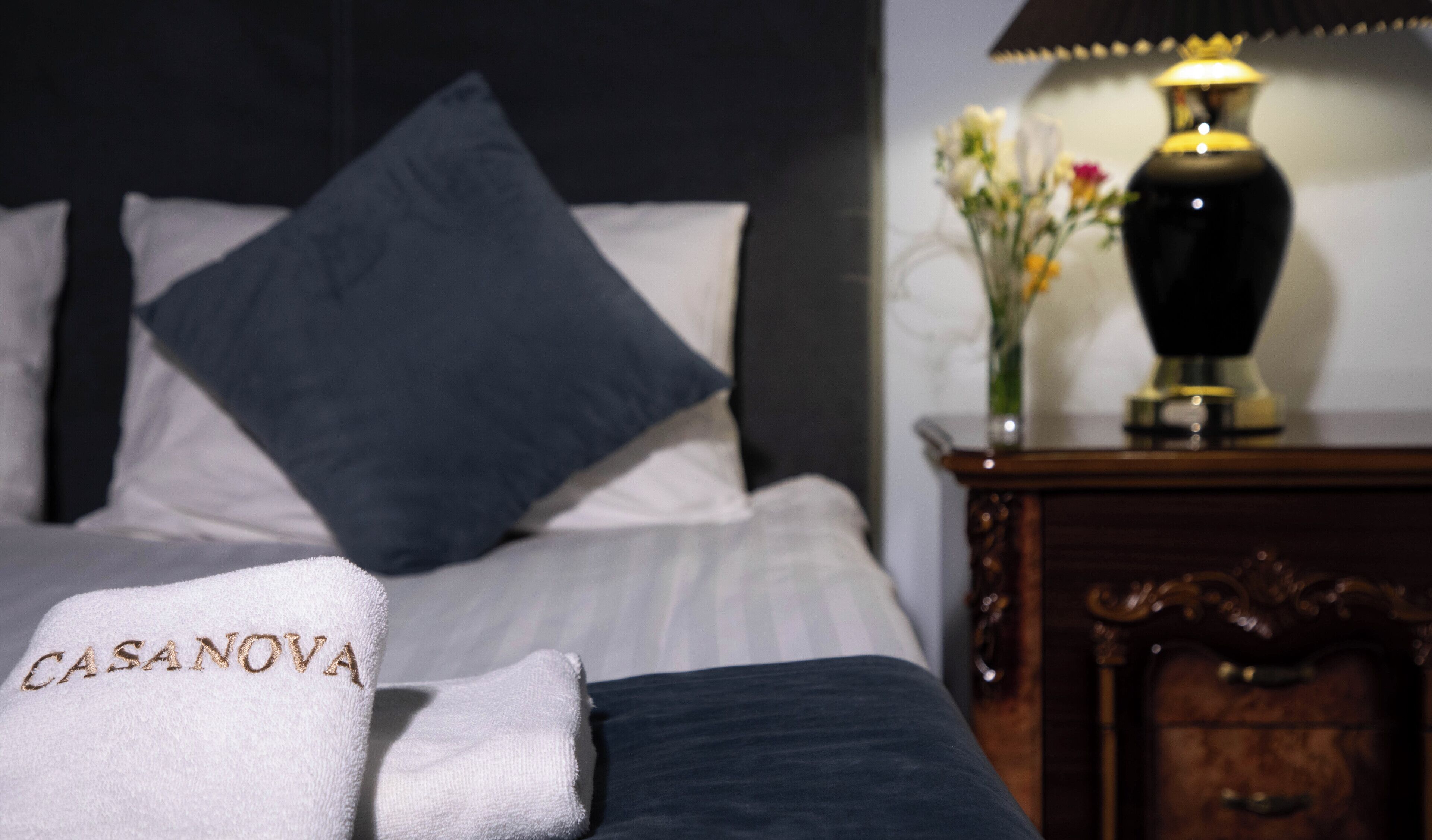 A neatly made bed with a white and dark blue pillow arrangement, a bedside table with a lamp and a vase of flowers, and a towel with "CASANOVA" embroidered on it.