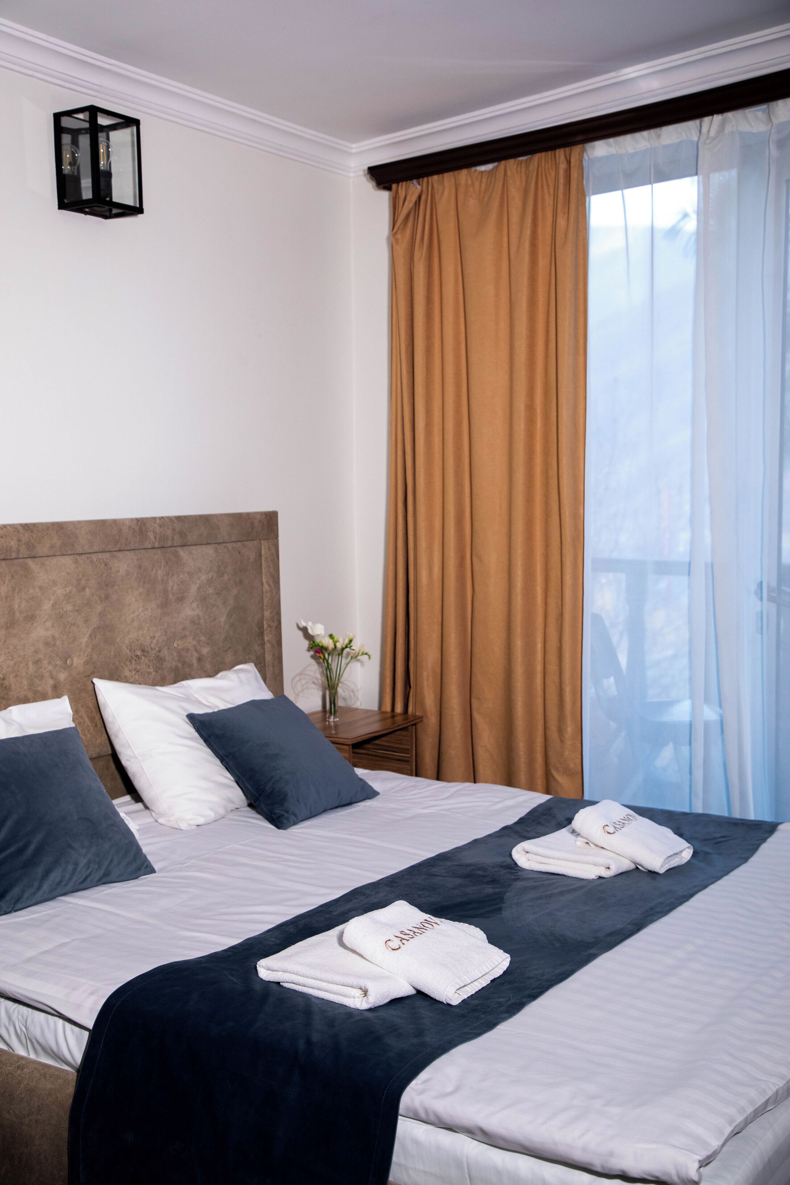 Una cama bien hecha con almohadas blancas y azul oscuro, una colcha azul oscuro y dos toallas dobladas con la palabra 'CASAMATOS' en ellas. Junto a la cama hay una pequeña mesa con un florero de flores.