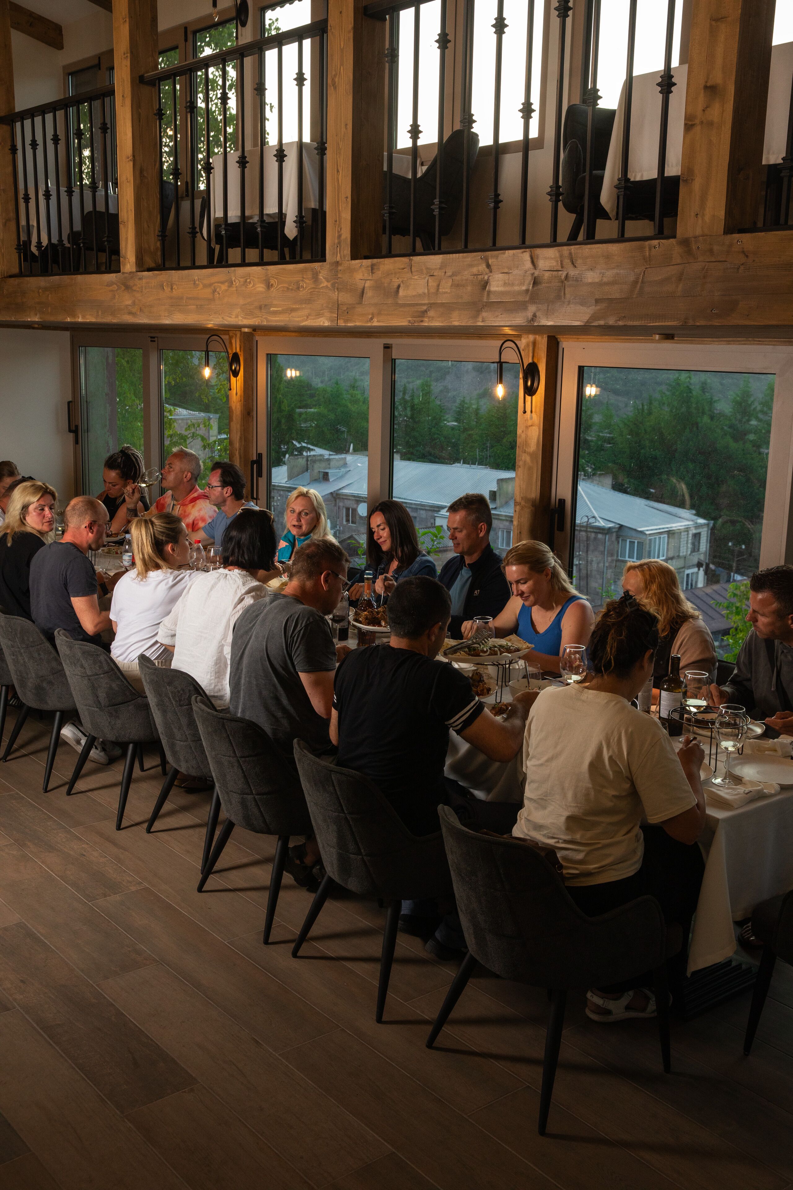 A restaurant with a high ceiling and wooden beams, large windows offering a view of the outdoors, and a group of people dining at a long table.