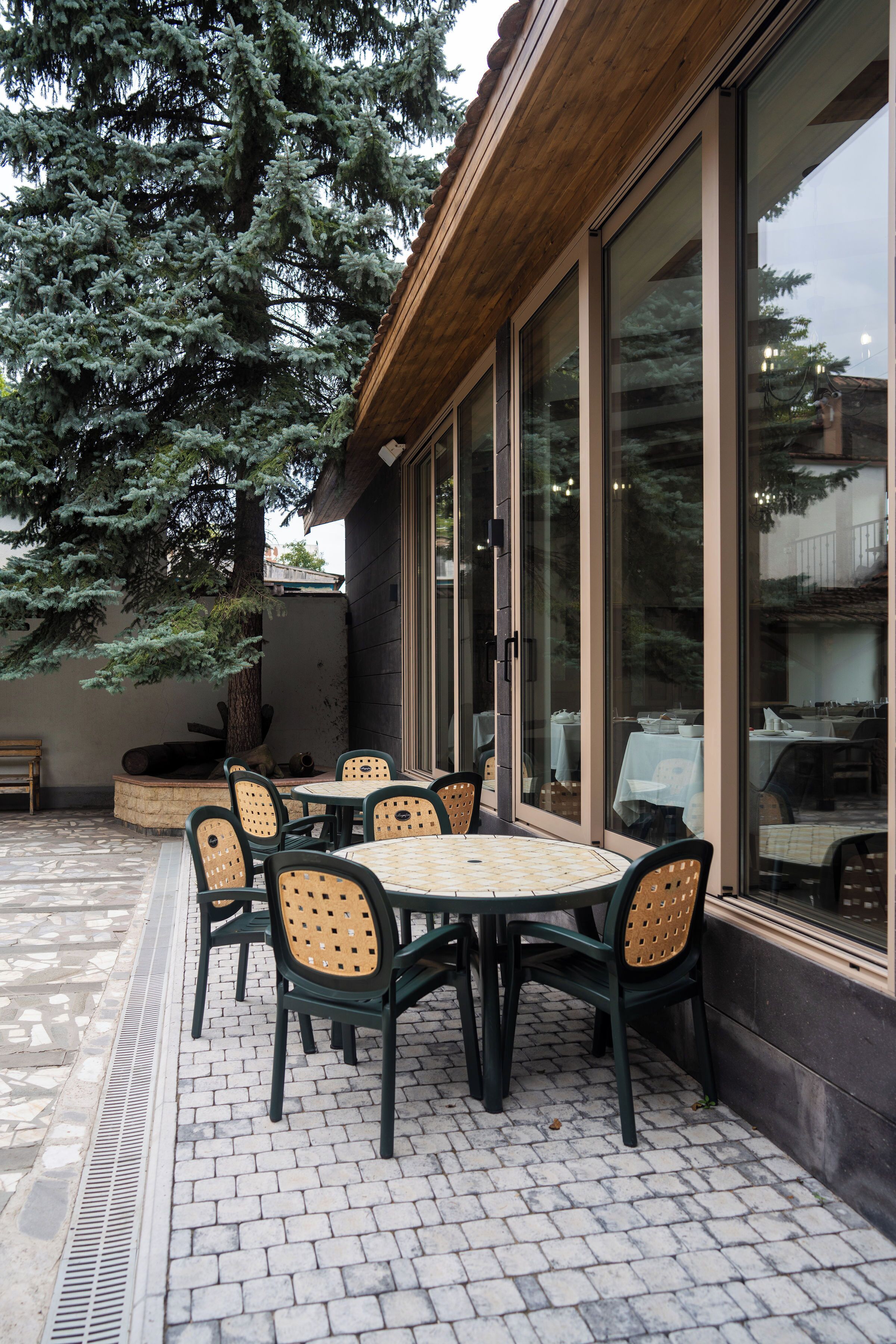 Un patio al aire libre con mesas y sillas redondas, ventanas amplias y un árbol.