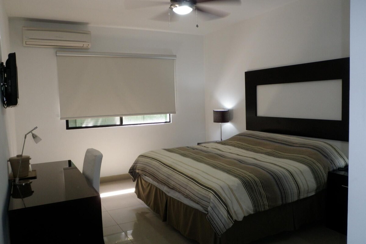 Ein Schlafzimmer mit Bett, Schreibtisch, Stuhl, Deckenventilator, einem Fenster mit Jalousien und einer Wand-Klimaanlage.