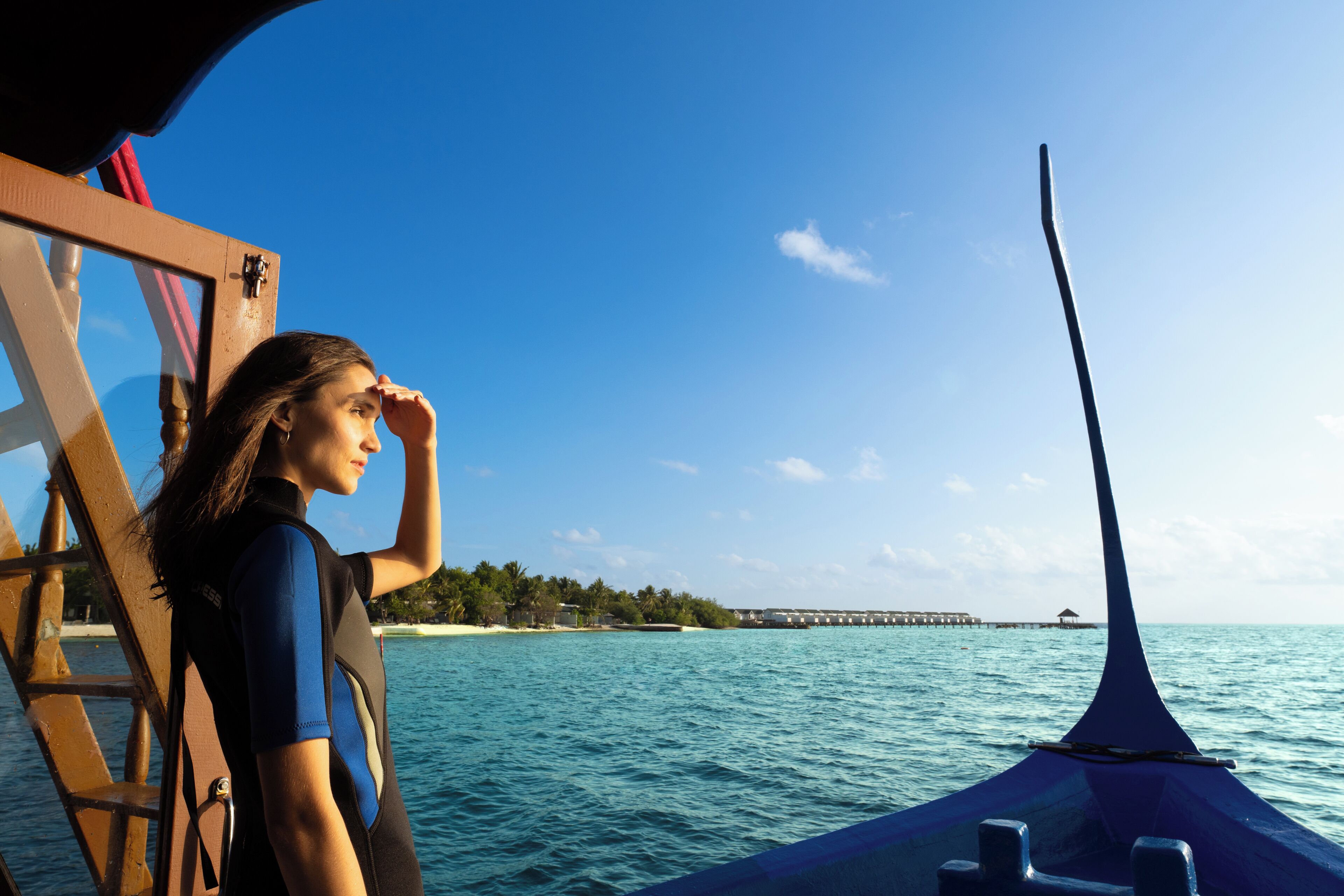 Uma pessoa com fato de mergulho em um barco, com vista para o mar e um cais ao longe.