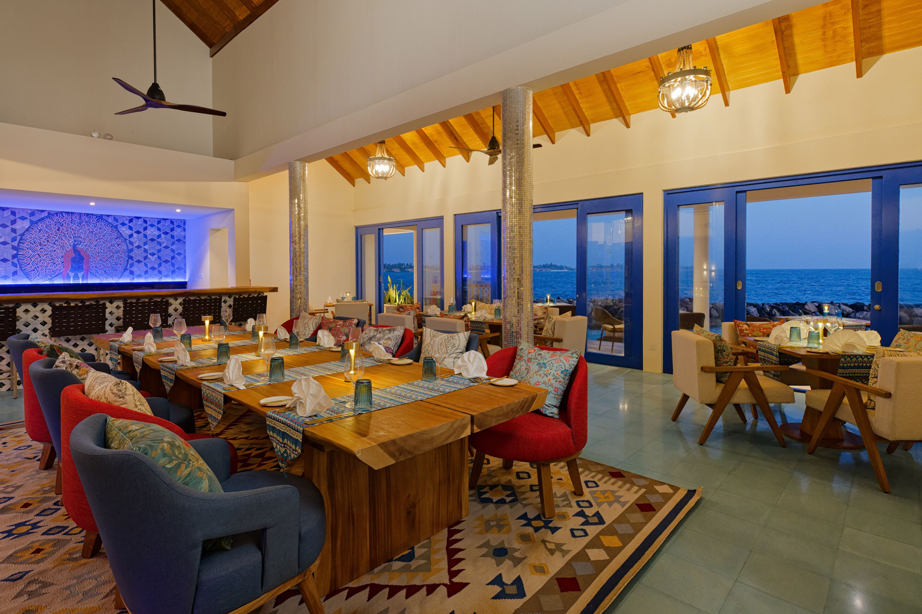 A dining area with a large wooden table, multiple chairs, and a view of the sea through large windows.