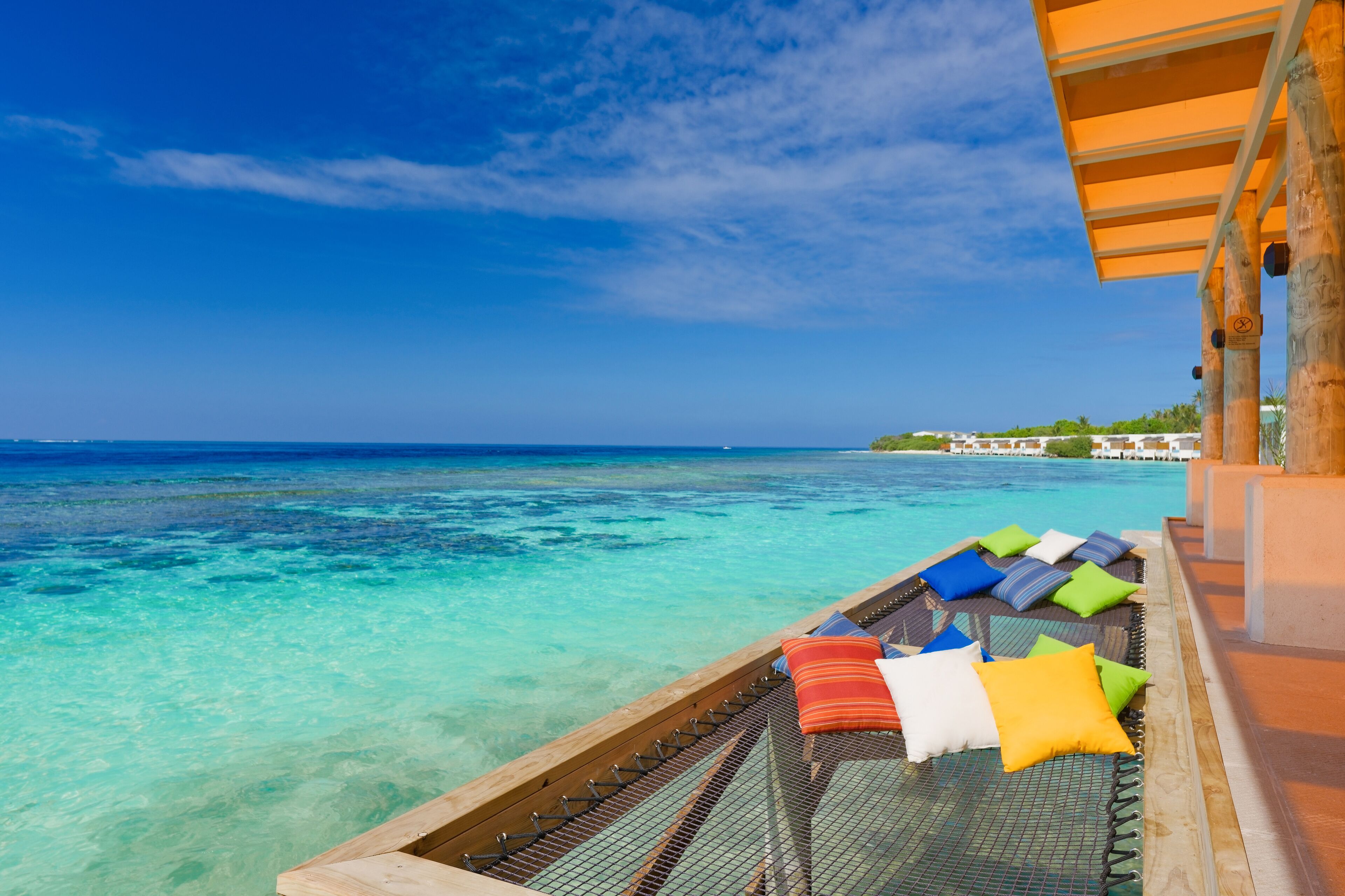 A beach view with a hammock and colorful pillows, clear turquoise water, and a wooden structure.