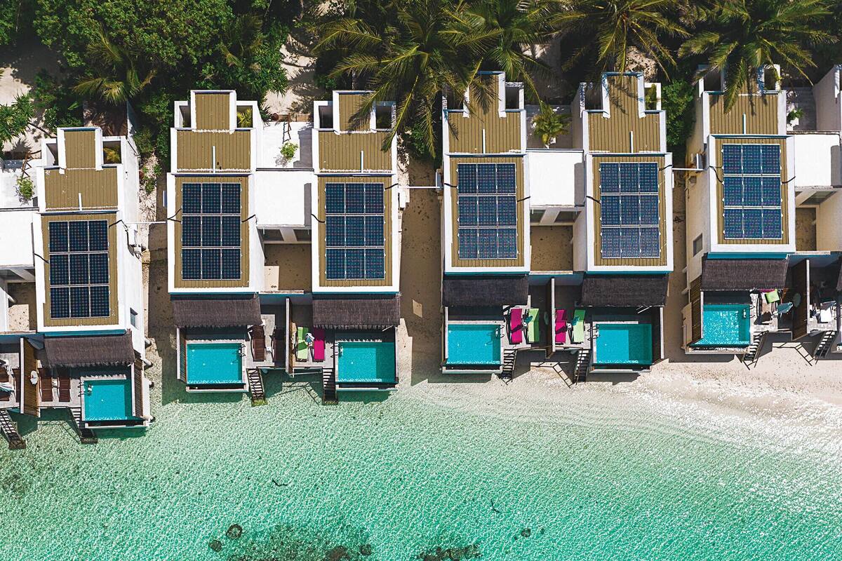 Un hôtel situé en bord de mer, doté de plusieurs bungalows sur pilotis, d’une eau turquoise cristalline et de palmiers.