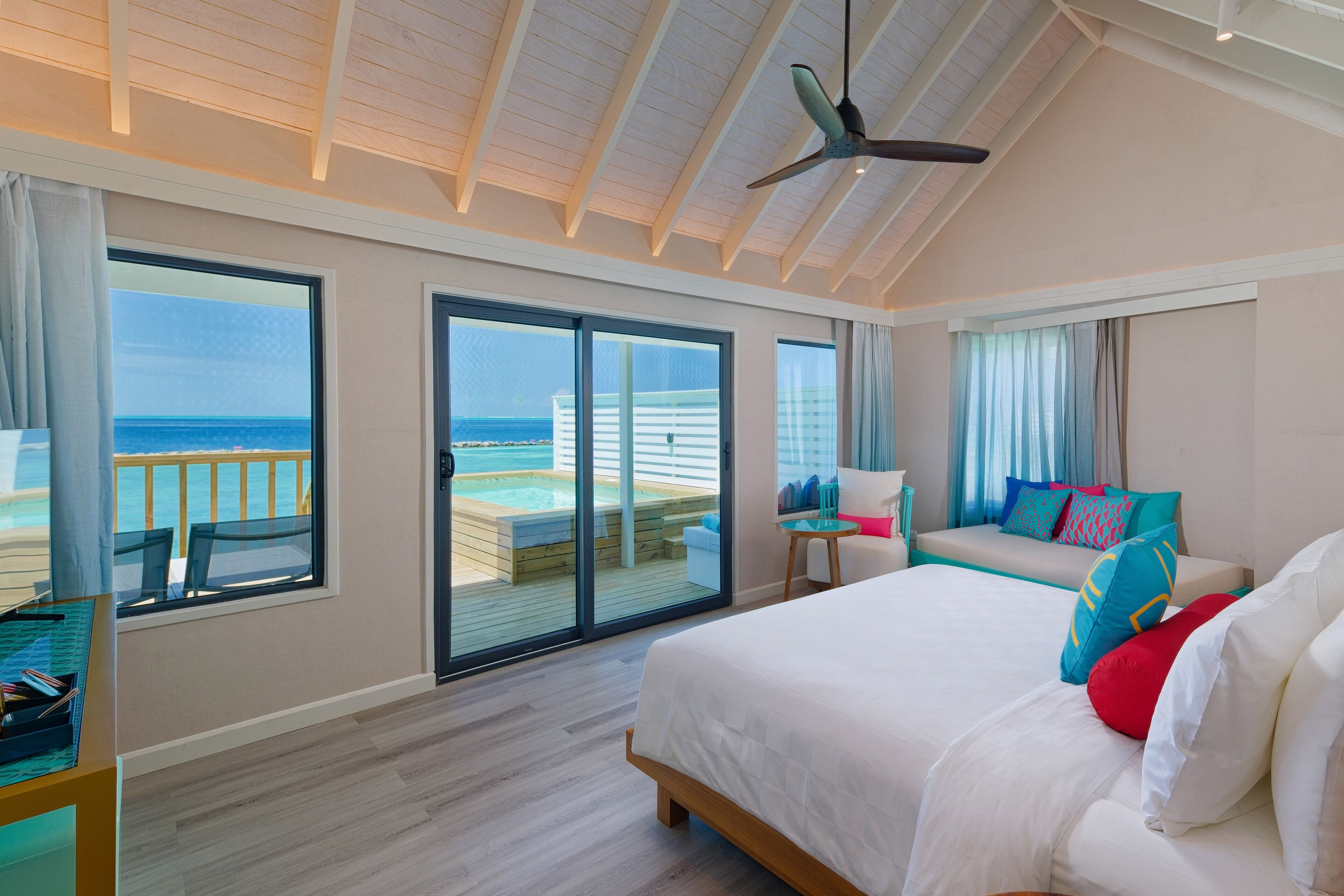 Un dormitorio con una cama grande, un ventilador de techo y vistas a la playa a través de amplias ventanas.