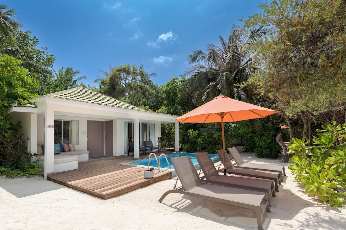Un espace aménagé au bord de la piscine, avec des chaises longues, une terrasse en bois et un bâtiment doté d’un toit recouvert de tuiles.
