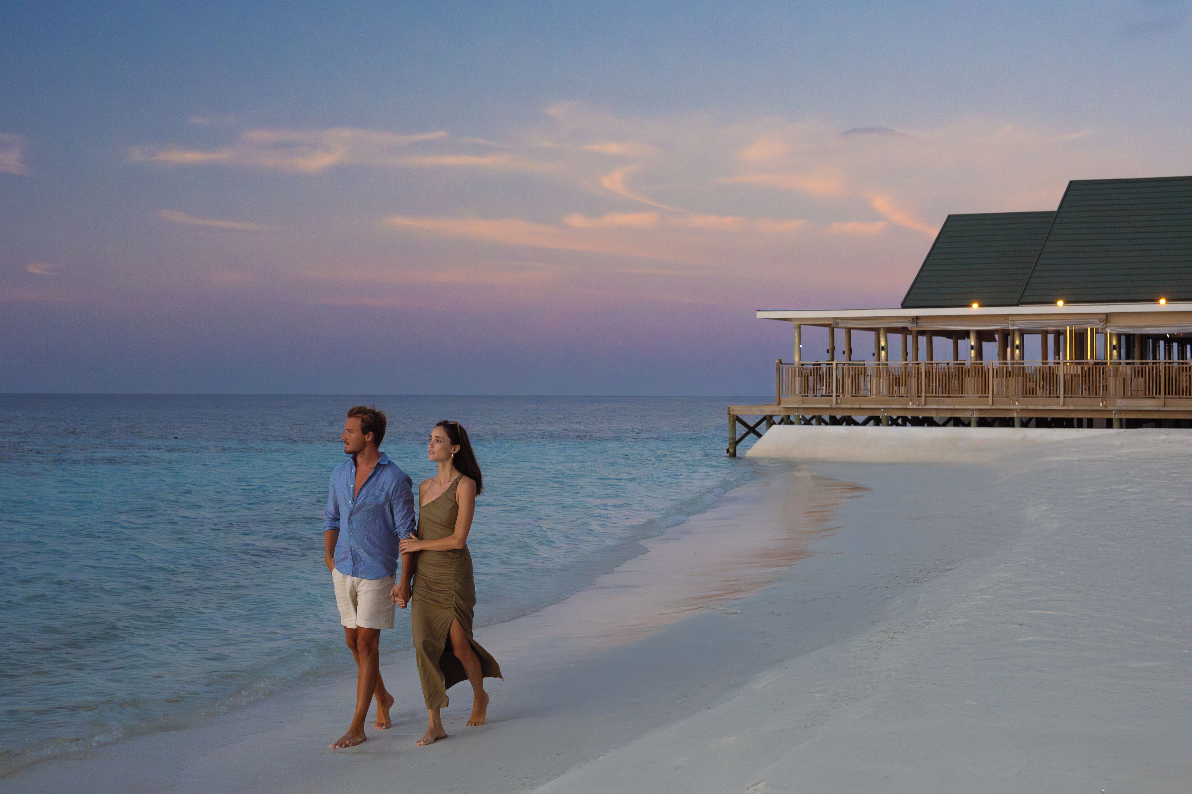 Una pareja caminando por una playa, con una estructura de madera al fondo.