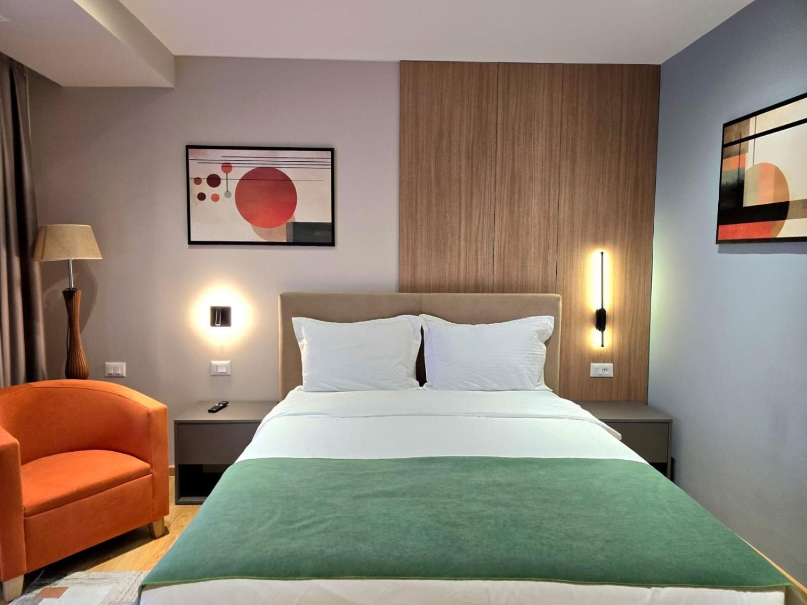 Habitació d'hotel amb un llit, una tauleta de nit, una làmpada, una cadira i obres d'art a les parets.