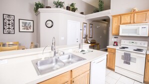 A modern kitchen with a double sink, wooden cabinets, a microwave, and an oven. There are plants on the upper cabinets and a clock on the wall. - Glenbrook Homes (Clermont)