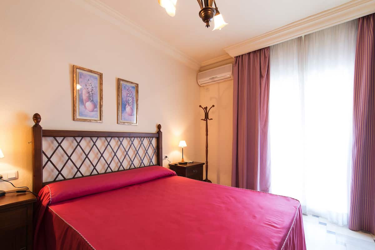 Hotelkamer met een bed, twee wanddecoraties, een airconditioningapparaat, een vloerlamp en een raam met gordijnen.
