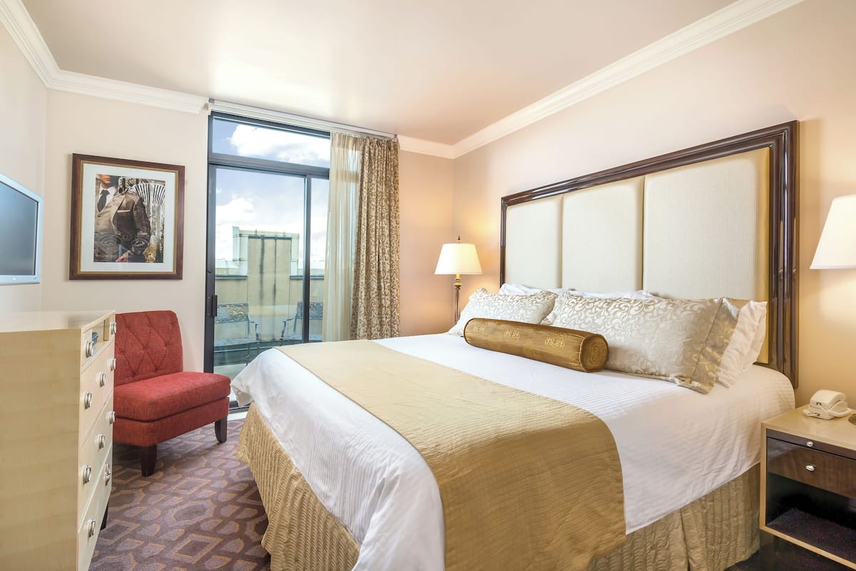 A hotel room with a large bed, a red chair, a desk with a lamp, a framed picture on the wall, and a view of the sky through a window.