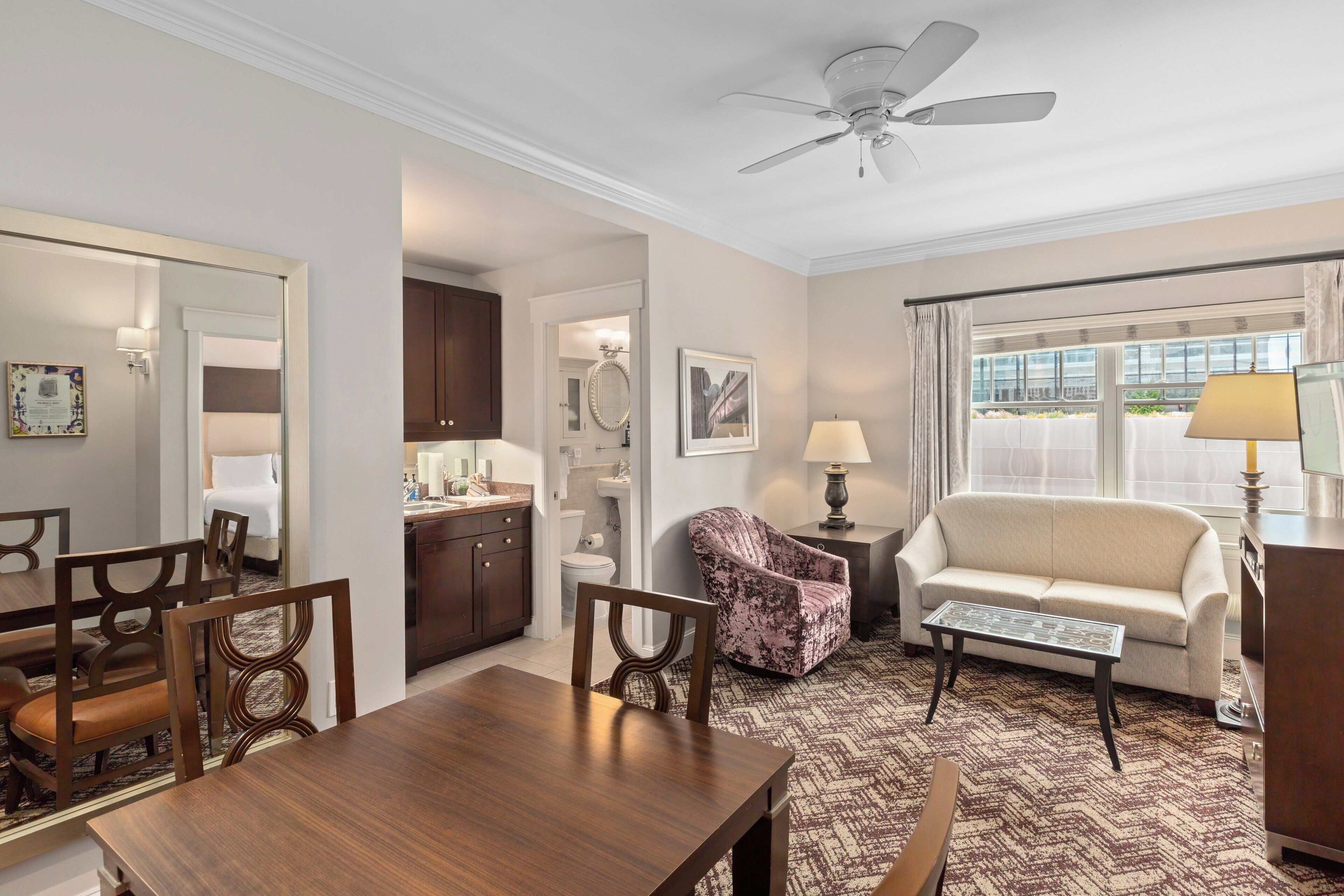 A hotel room with a dining area, a sofa, a chair, a table, a ceiling fan, and a window with curtains.