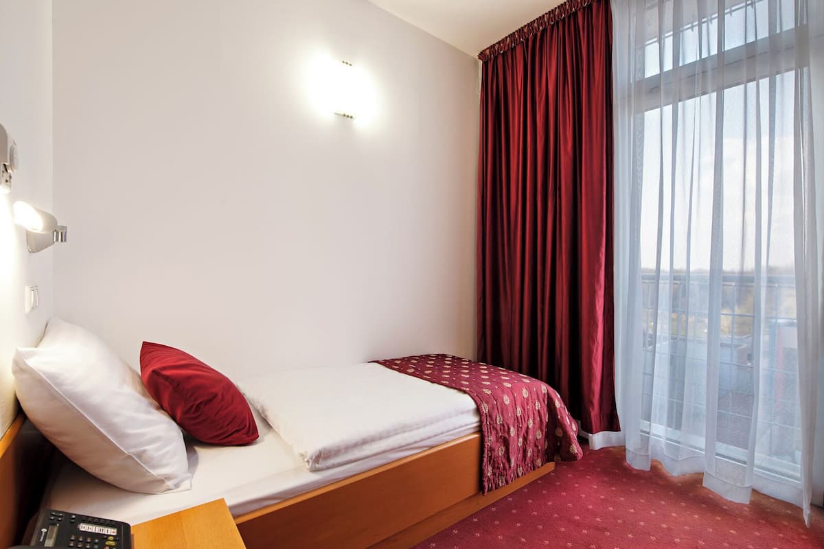 A single bed with a red and white bedspread, a red pillow, and a wooden headboard. There is a remote control on a wooden nightstand beside the bed. The room has a large window with sheer white curtains and a view of the outside.