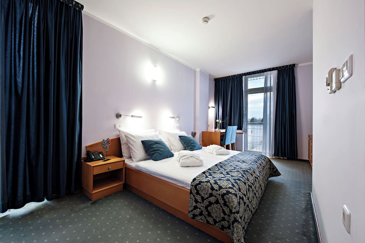 A hotel room with a bed, bedside table, lamp, and a view of the outdoors.