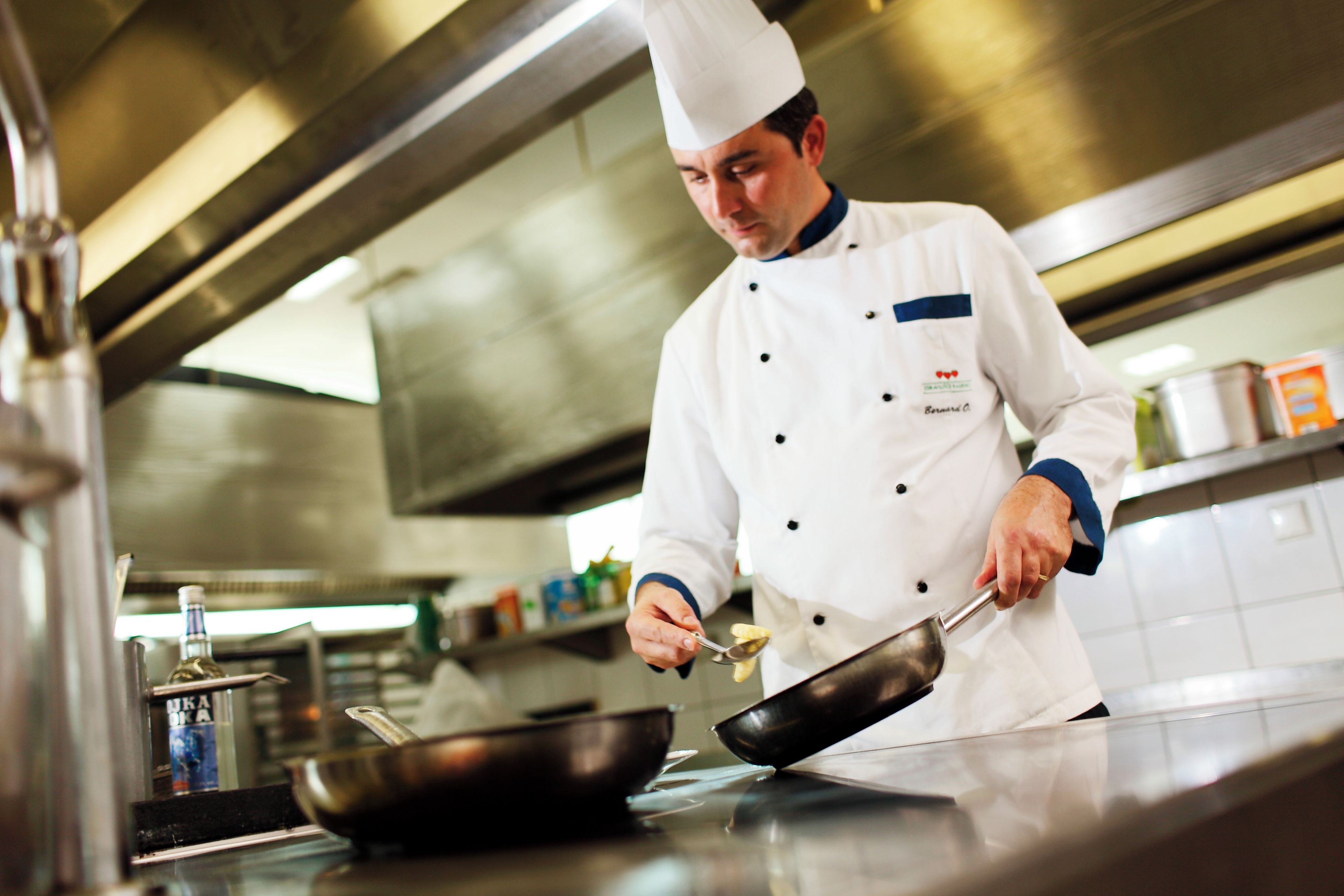 A chef in a professional kitchen environment, wearing a chef's uniform and hat, with stainless steel appliances and cookware.