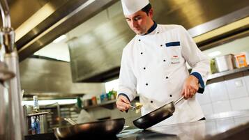 A chef in a professional kitchen environment, wearing a chef's uniform and hat, with stainless steel appliances and cookware.