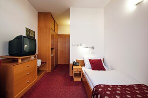 A hotel room with a wooden wardrobe, a television on a wooden stand, a bed with white and red bedding, a nightstand with a lamp, and a red carpet.