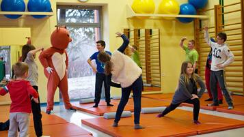 A group of children and an adult in a room with orange mats, a clock on the wall, and balloons.