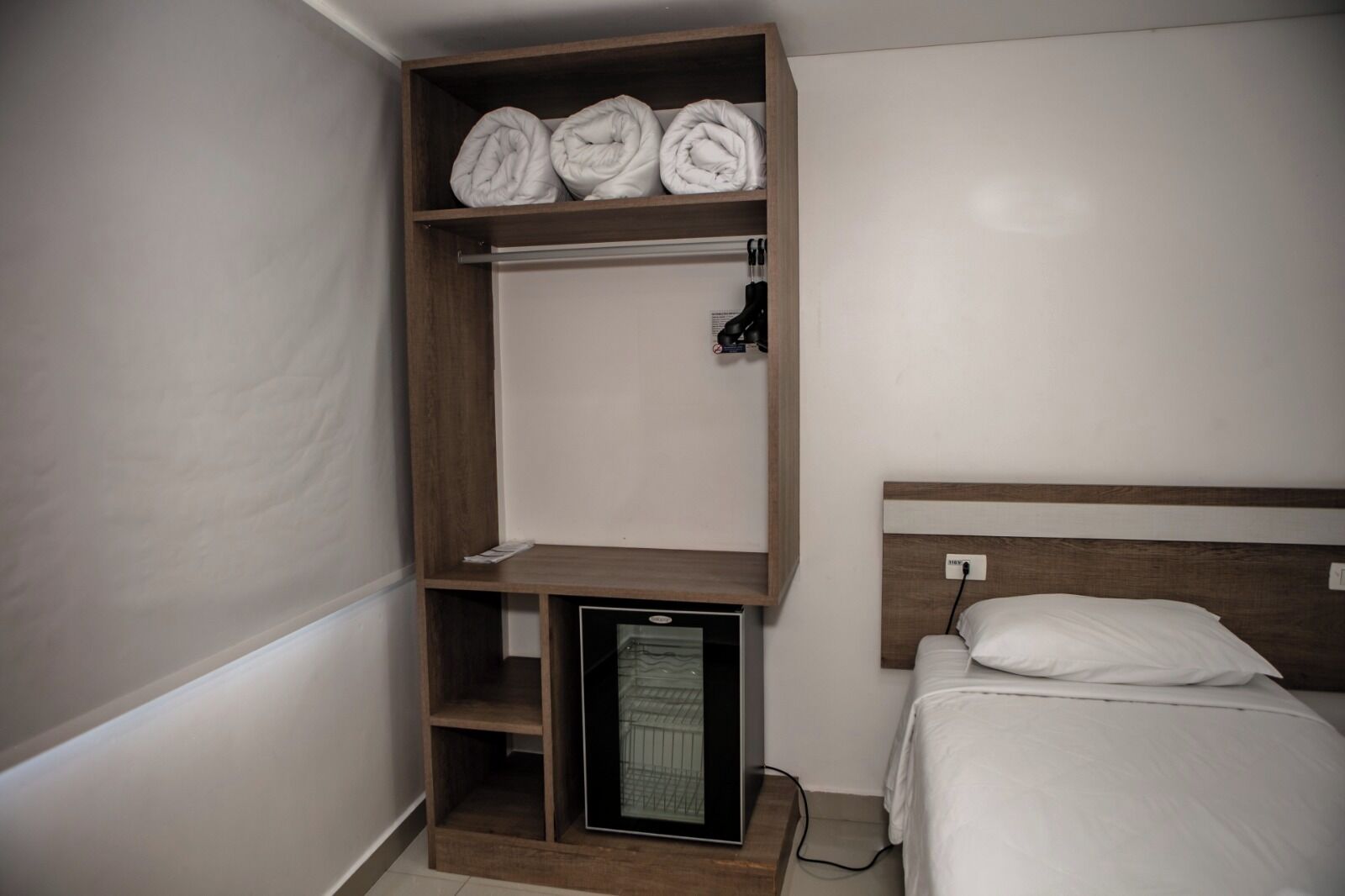 Quarto com cama, prateleira pequena com toalhas dobradas, mini-geladeira e tomada na parede.