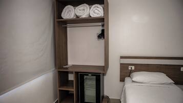 Quarto com cama, prateleira pequena com toalhas dobradas, mini-geladeira e tomada na parede.