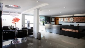 A modern reception area with a reception desk, seating area, and decorative elements like a chandelier and a red plant. - San Lucas Hotel (Sarandi)