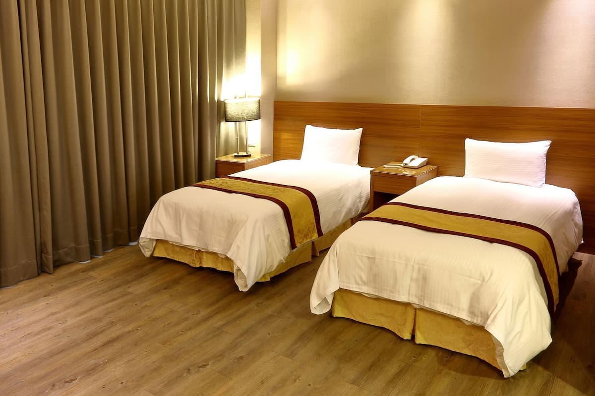 A hotel room with two beds, a wooden headboard, a nightstand with a lamp, and a curtain.