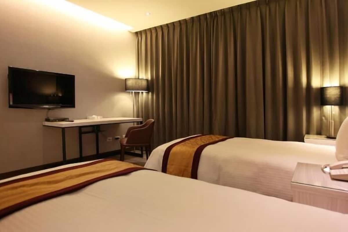 A hotel room with two beds, a desk, a chair, a television, and a lamp.