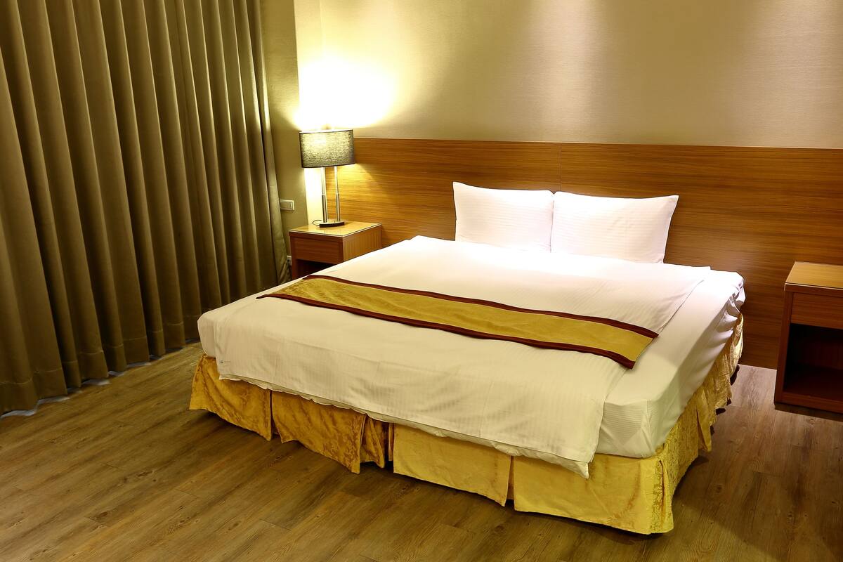 A hotel room with a neatly made bed, wooden headboard, and a nightstand with a lamp.