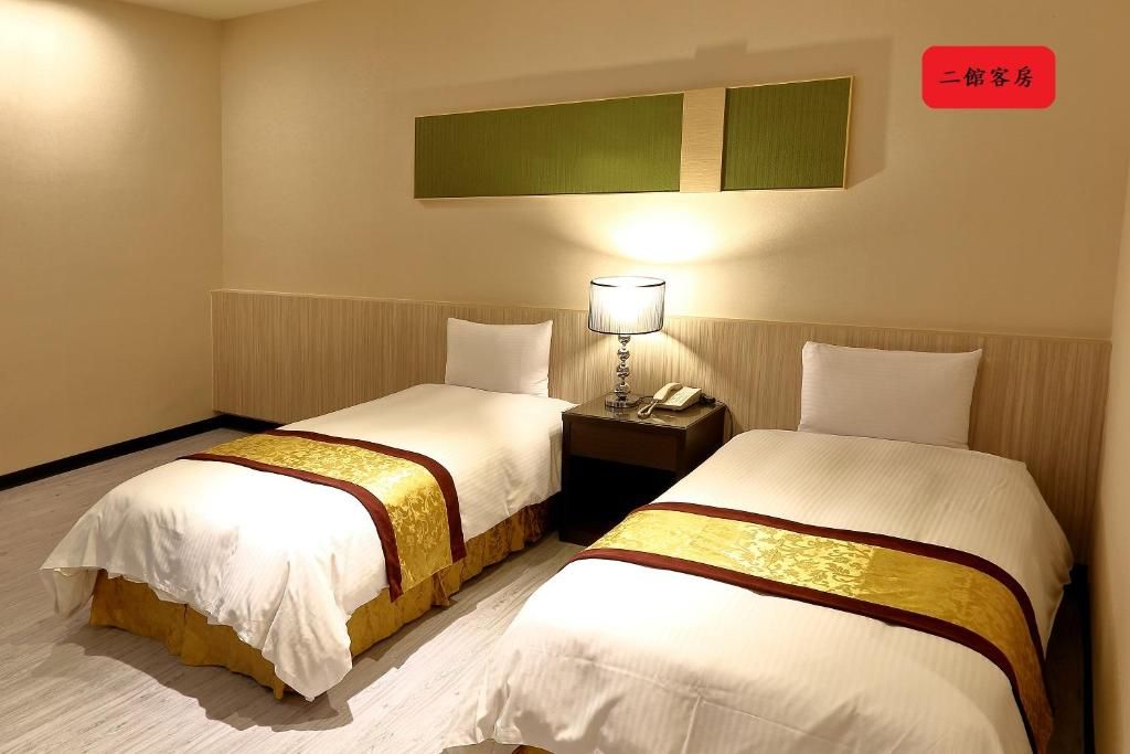 A hotel room with two beds, a nightstand with a lamp, and a wall-mounted panel.