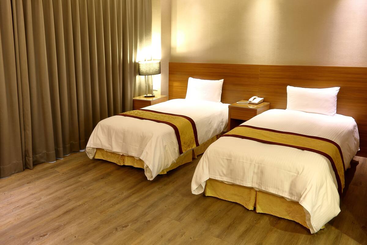 A hotel room with two beds, wooden headboard, a nightstand with a lamp, and a telephone.